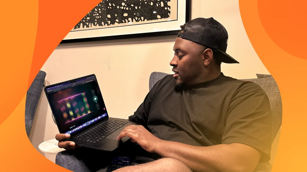 A man in casual clothes, sitting on a chair and holding a laptop playing progressive jackpots at SlotsLV, wearing a backwards cap and looking at the screen intently.